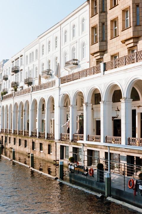 A white building with arched columns and balconies stands next to a body of water, with a beige building visible on the right.