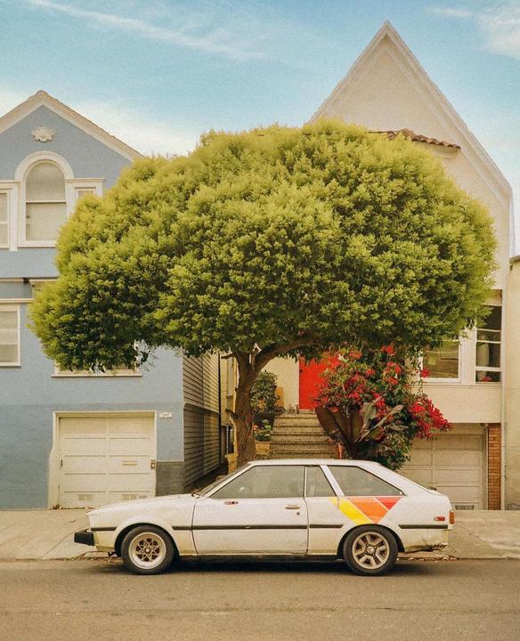 A white car with a colorful stripe is parked on a street in front of a house with a large tree.