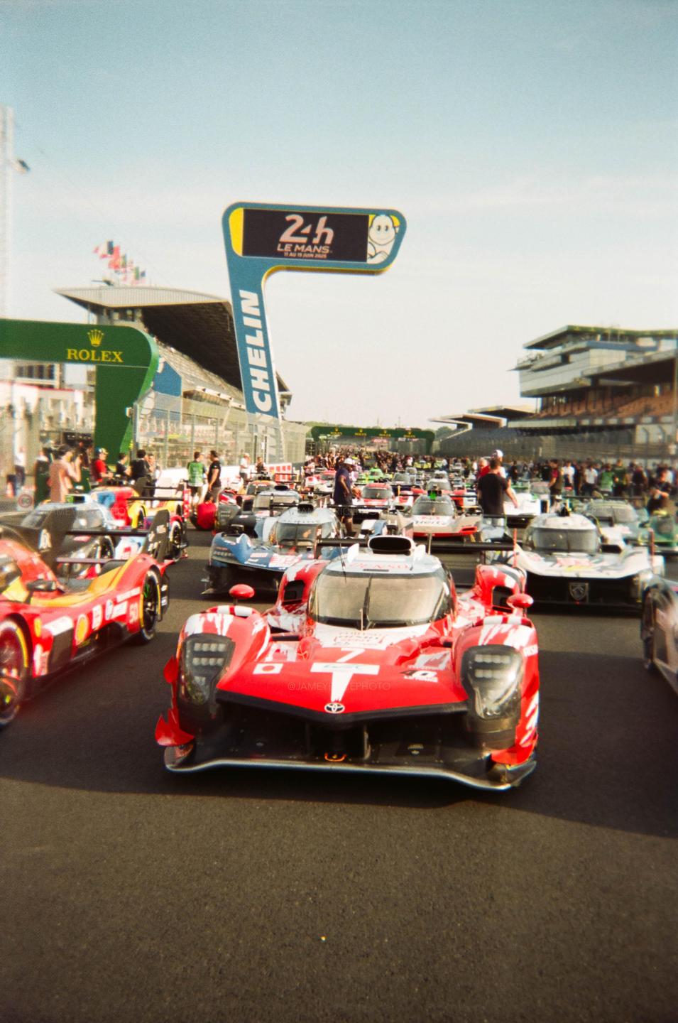 A red race car is in the foreground of a line of race cars on a track with a "24h Le Mans" sign in the background.