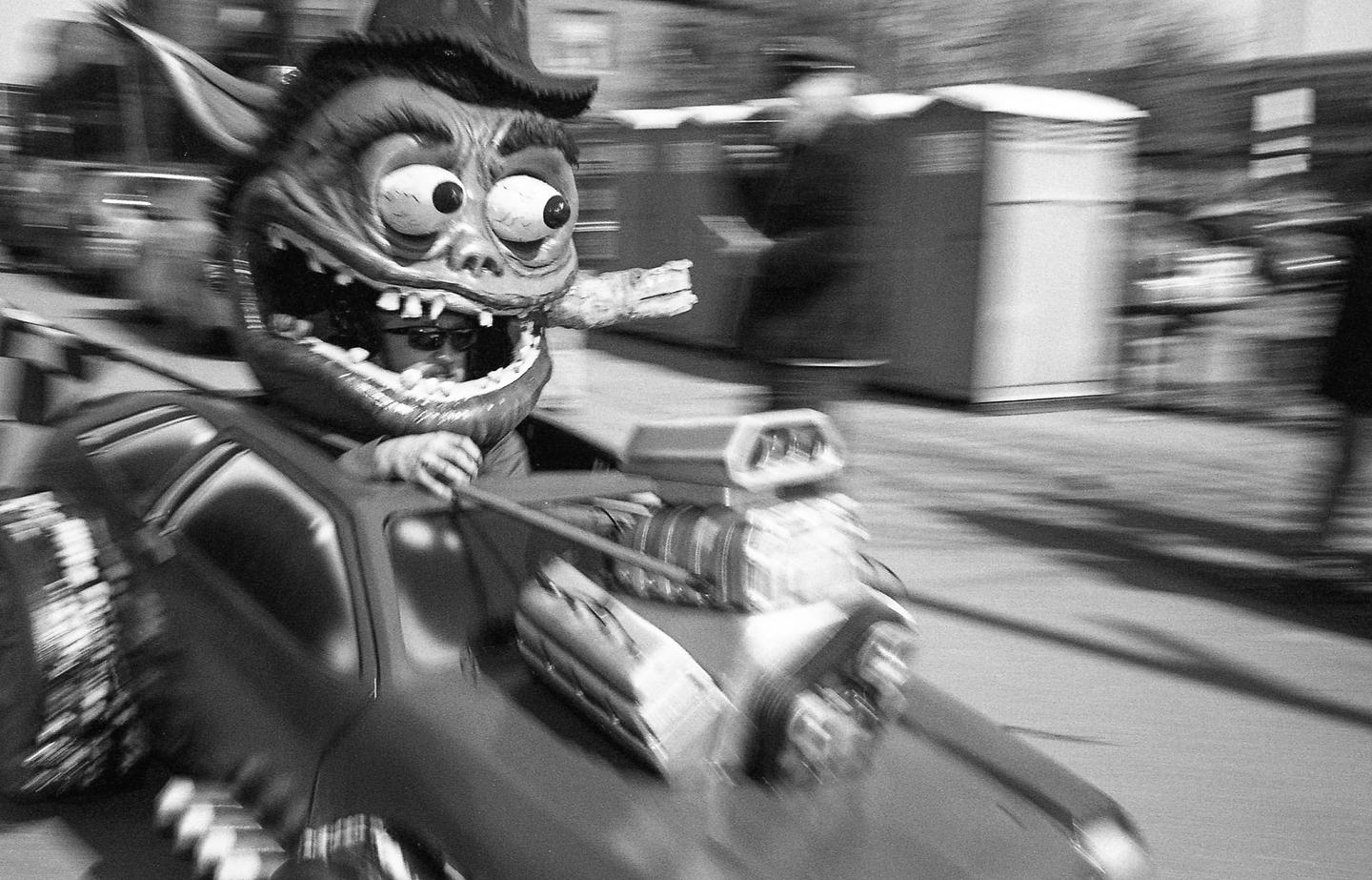 A person wearing a monster mask and sunglasses drives a toy car with a blurred background.