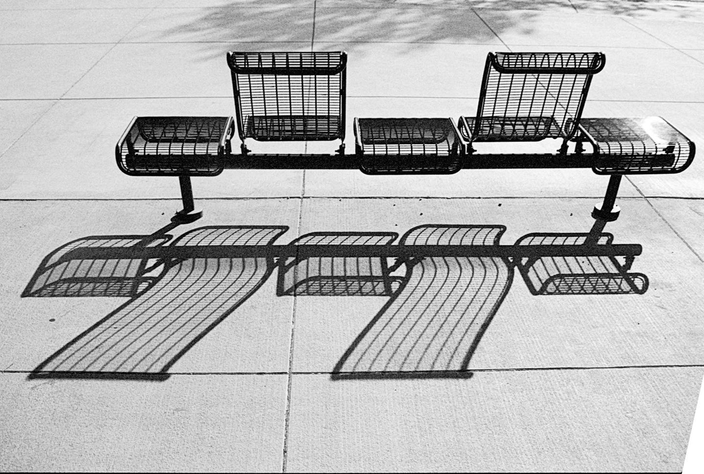 A black and white photo shows a metal bench casting a long shadow on a concrete surface.