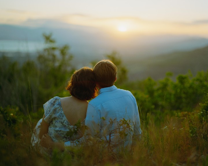 A couple sits in tall grass, looking out at a sunset over a mountain range.