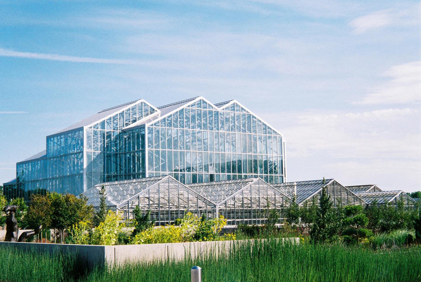 A large glass greenhouse building with a stepped roofline is surrounded by smaller greenhouses and lush green landscaping under a bright blue sky.