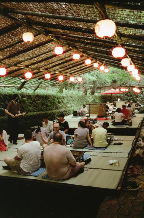 People sit on mats under a bamboo roof with many red lanterns, with a server carrying a tray on the left.