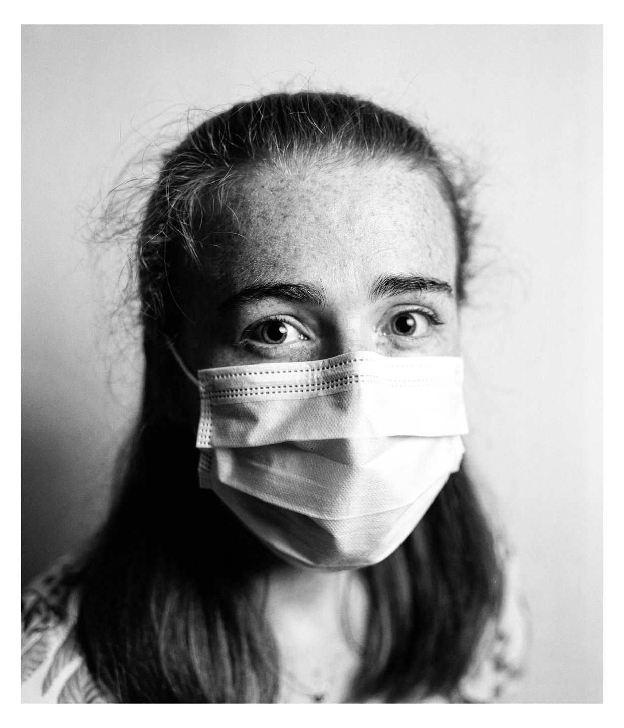 A young woman with freckles and long hair wears a surgical mask.