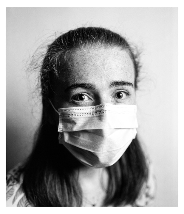 A young woman with freckles and long hair wears a surgical mask.