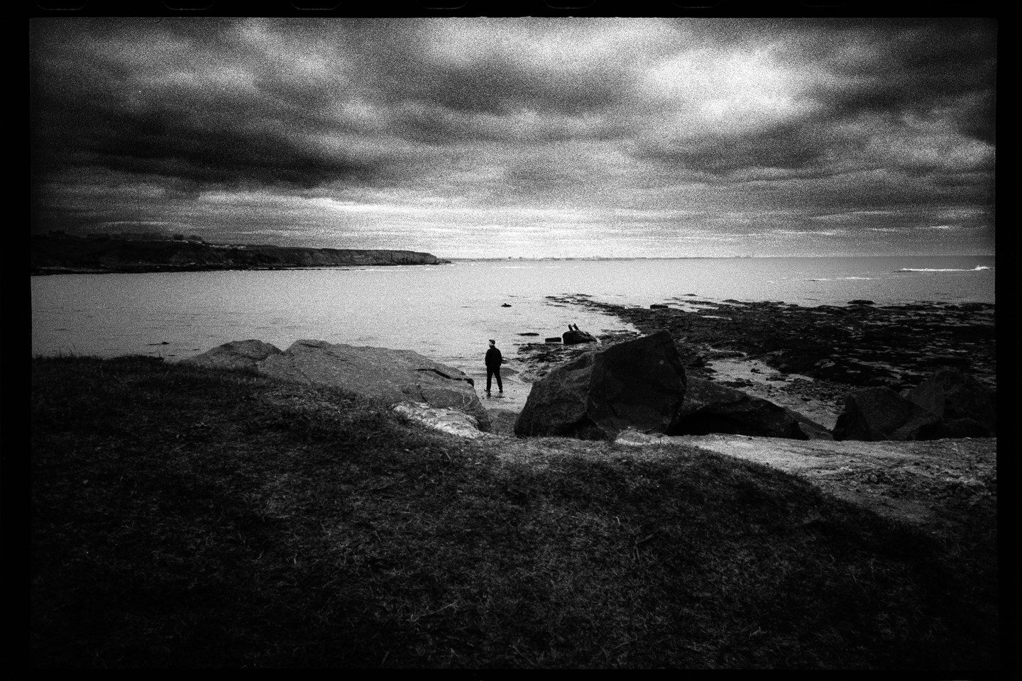 A person stands on a rocky beach under a dramatic, cloudy sky.