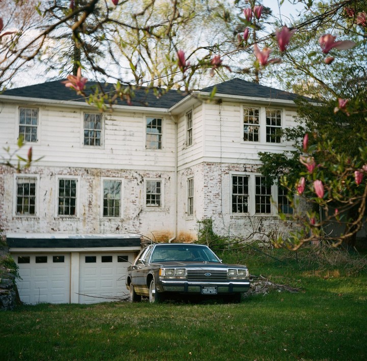 A dark sedan is parked on a grassy lawn in front of a white house with a two-car garage.
