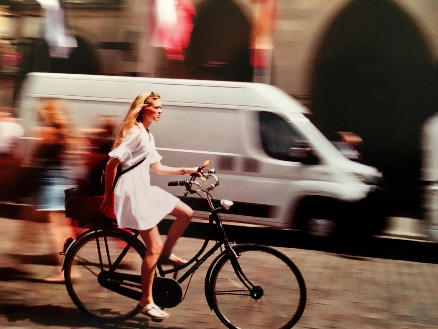 A woman in a white dress rides a bicycle past a blurred white van and other blurred figures on a sunny street.