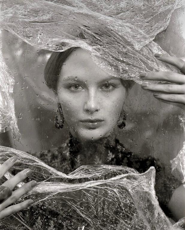 A woman with dark earrings looks directly forward through a plastic sheet covered in water droplets.