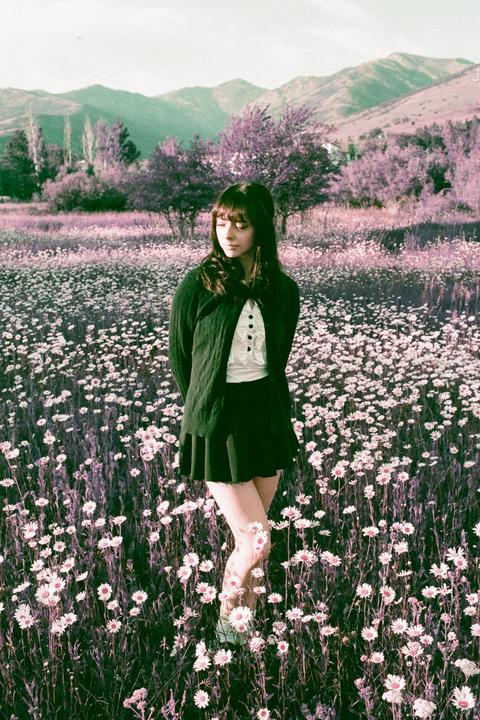 A woman stands in a field of white flowers with purple foliage and mountains in the background.