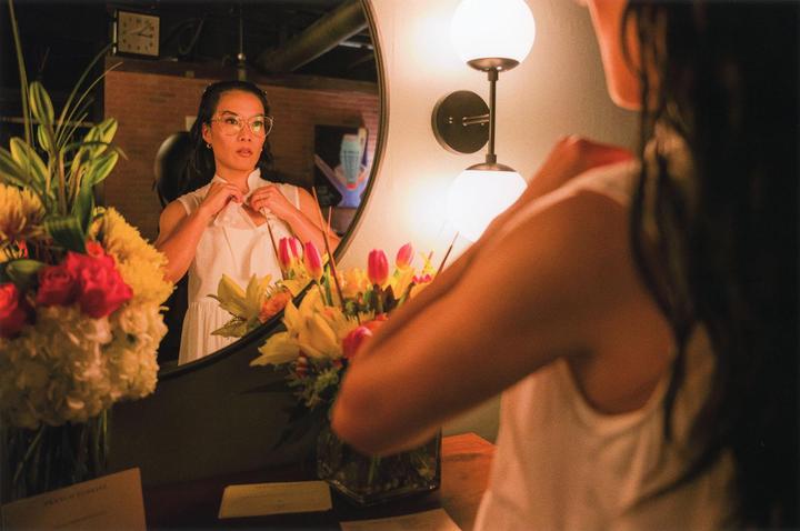 A woman in a white dress adjusts her collar while looking at her reflection in a mirror, with bouquets of flowers on either side.