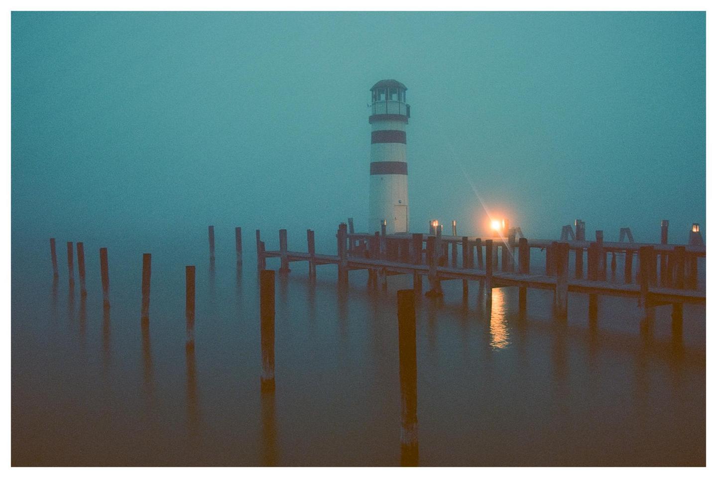 A striped lighthouse stands at the end of a pier on a foggy evening, with lights reflecting on the water.