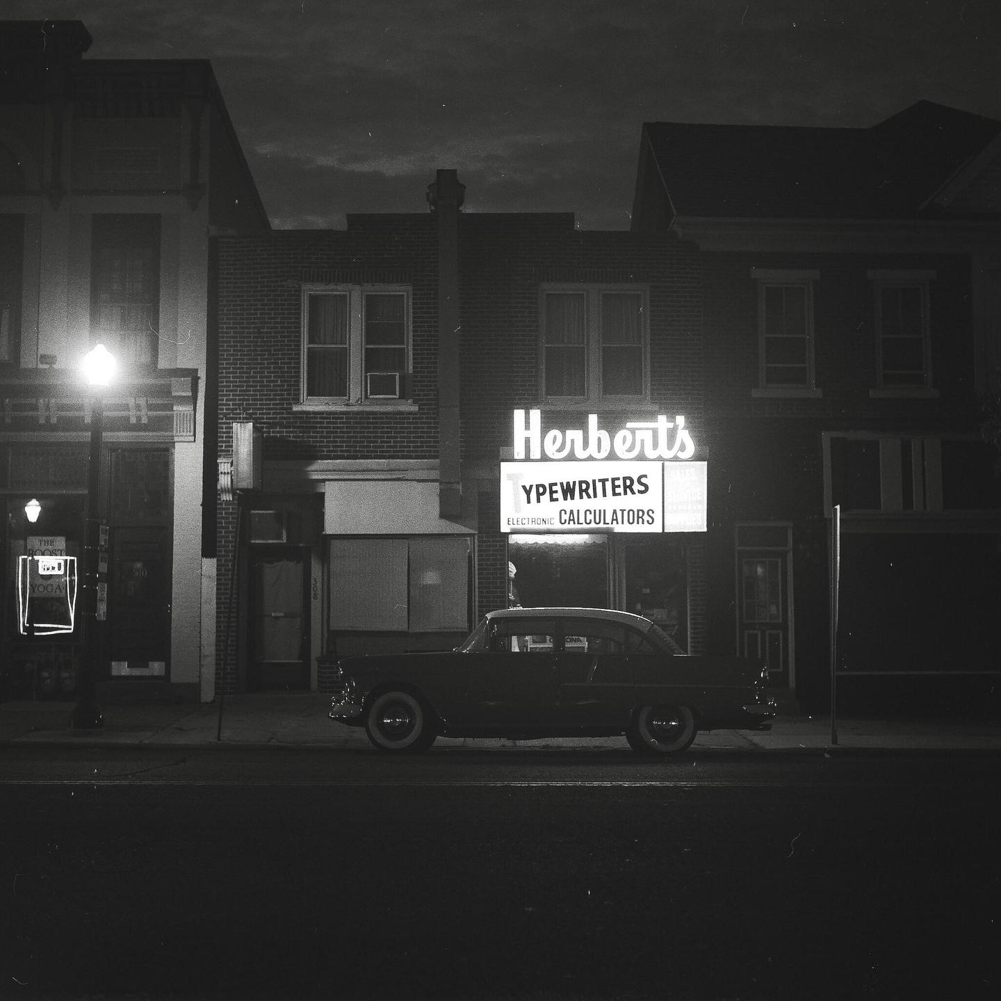 A vintage car is parked on a street in front of a building with a neon sign for Herbert's Typewriters and Calculators.