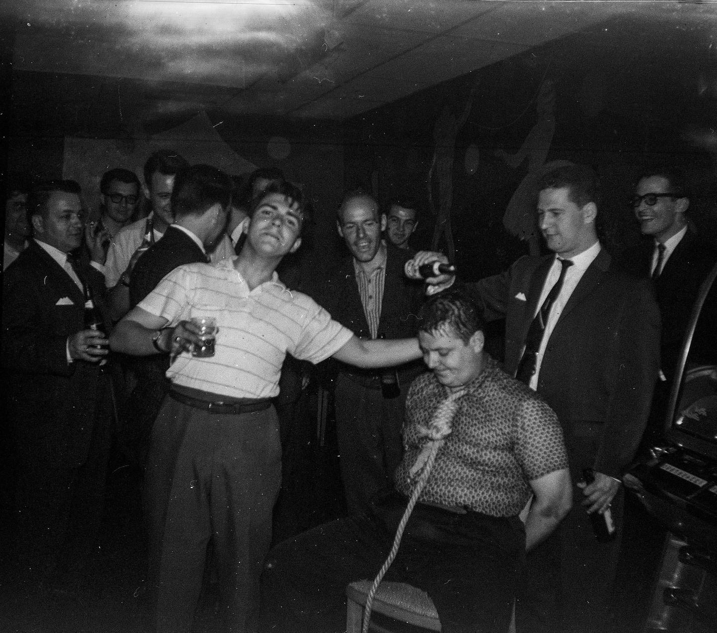 A group of men are gathered in a dimly lit room, with one man tied to a chair and another pouring a drink on his head.