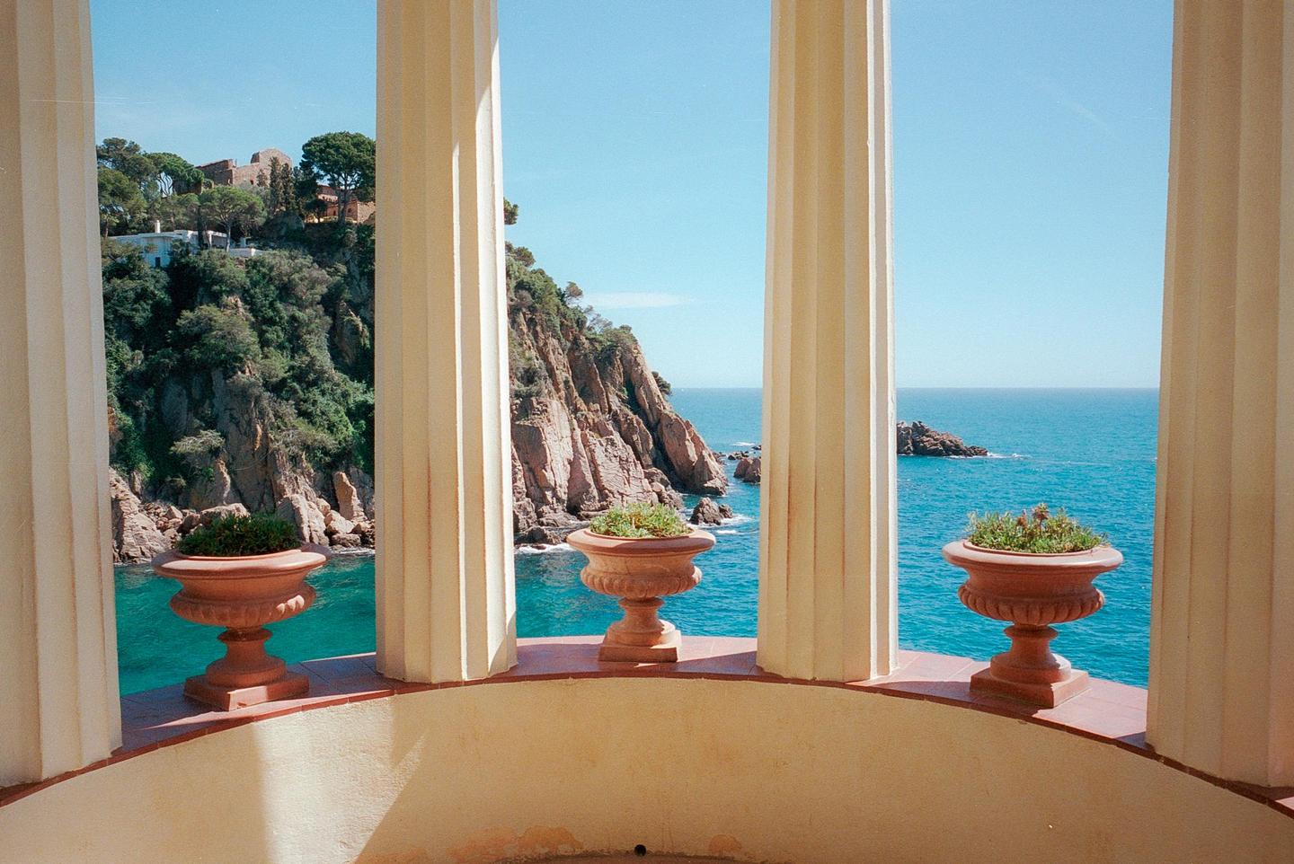 Three decorative planters sit on a balcony overlooking a rocky coastline and the sea, framed by architectural columns.