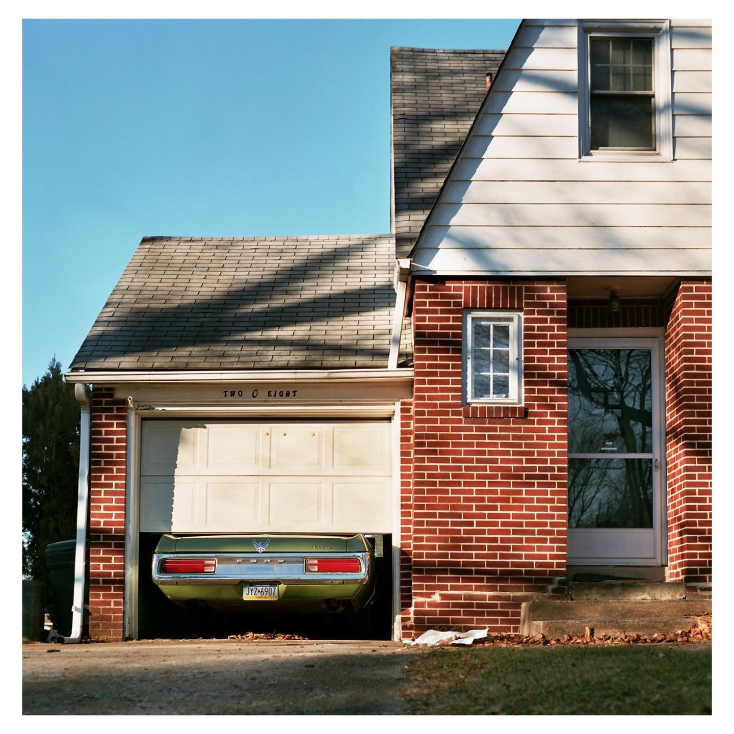 A green car is parked in a garage with the door partially open next to a brick house.