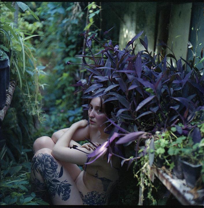 A woman with tattoos is partially obscured by purple foliage in a lush green setting.