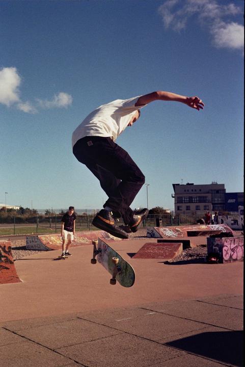 A man in a white t-shirt and dark pants jumps on a skateboard in a skatepark under a blue sky.
