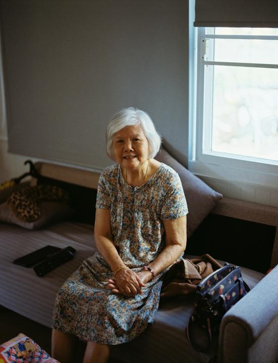An elderly woman with white hair sits on a couch next to a window.