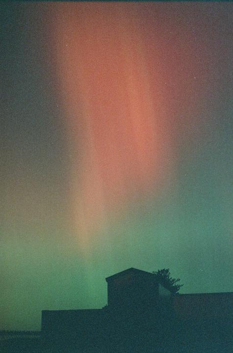 The northern lights glow in shades of red and green above a dark building and tree.