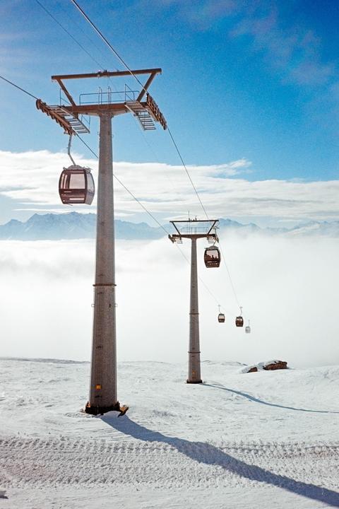 Gondolas ascend a snowy mountain with clouds and distant peaks in the background.