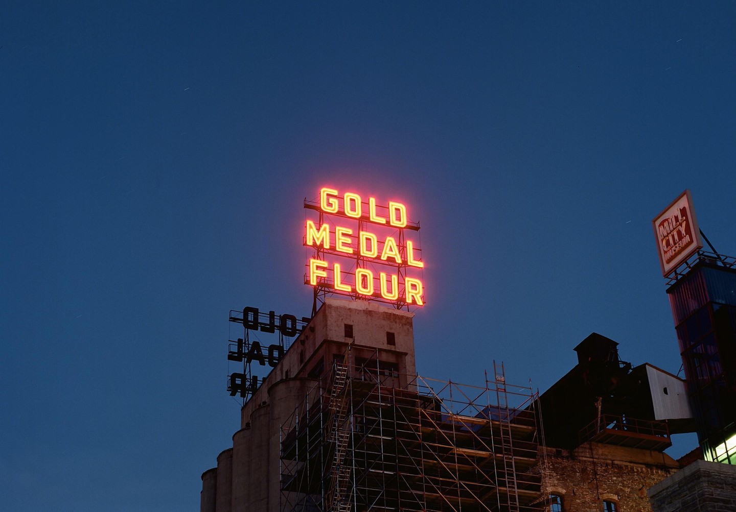 A neon sign for Gold Medal Flour sits atop a building with scaffolding against a dark blue sky.