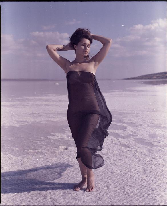 A woman in a sheer dark dress stands barefoot on a salt flat with her hands behind her head.