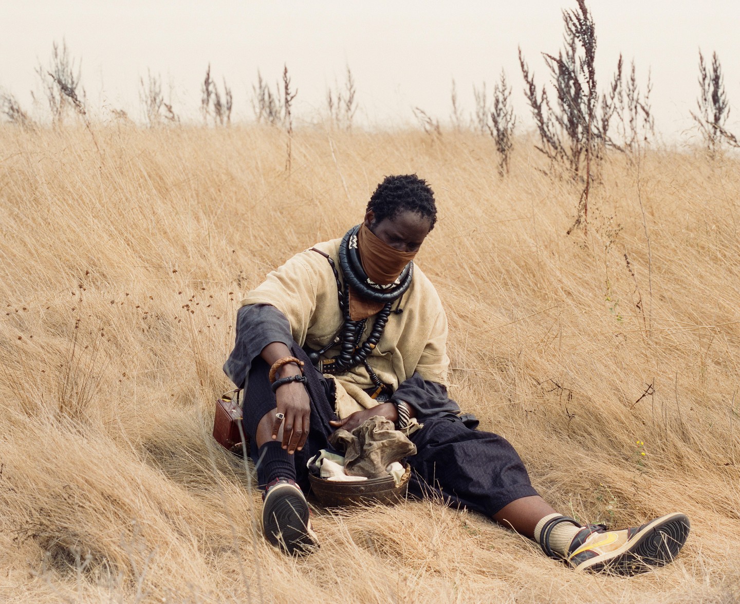 A person with a face covering and many necklaces sits in a field of tall, dry grass.