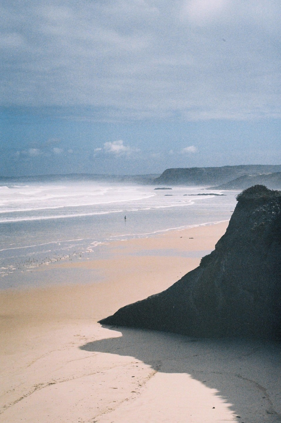 A lone figure stands in the shallow water of a vast beach with waves rolling in, framed by a dark cliff in the foreground and distant headlands under a cloudy sky.