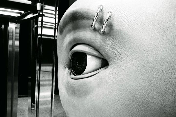A mannequin's eye with eyebrow piercings is visible next to a reflective surface.