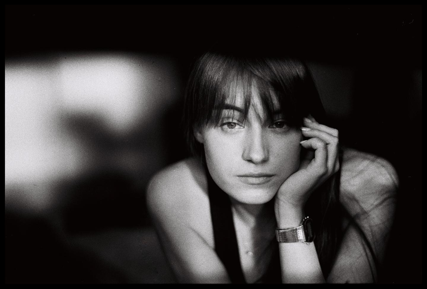A woman with bangs and a watch rests her chin on her hand while looking at the viewer.