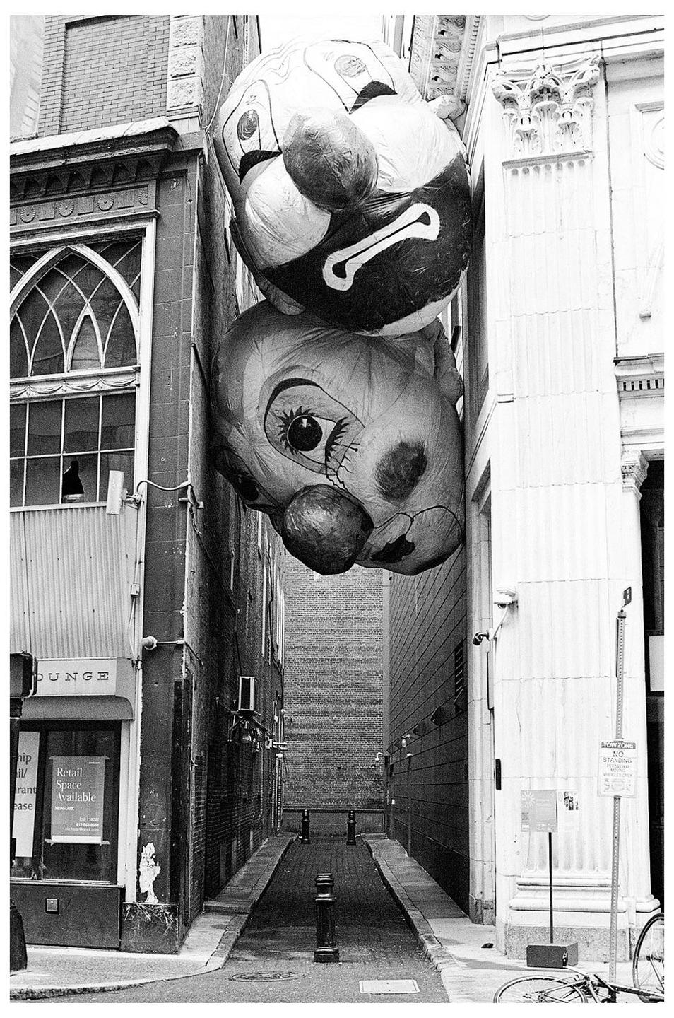 Two large clown balloons are wedged between two buildings on a narrow street.
