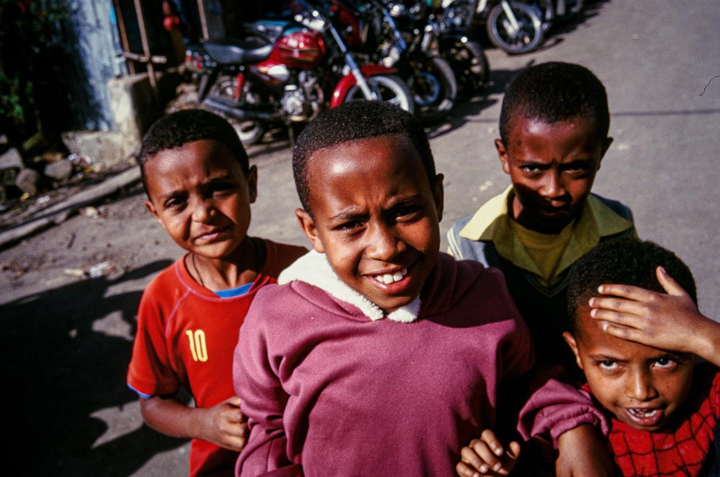 Four boys are standing on a street with motorcycles parked in the background.
