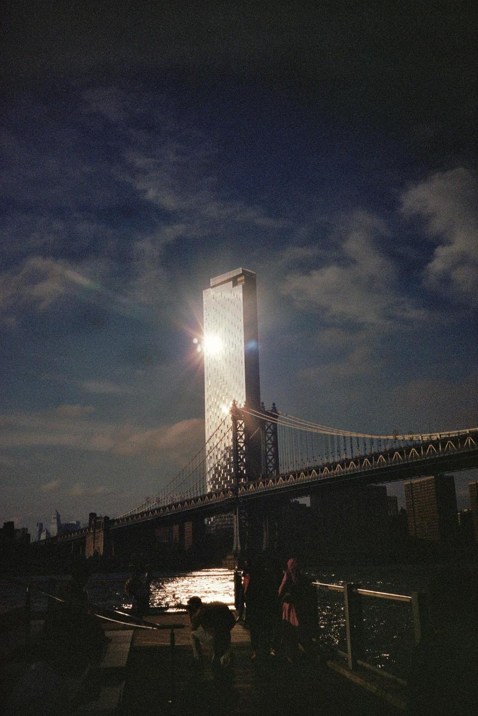 A tall building with sun glare on its facade stands next to a suspension bridge over water, with people silhouetted in the foreground.