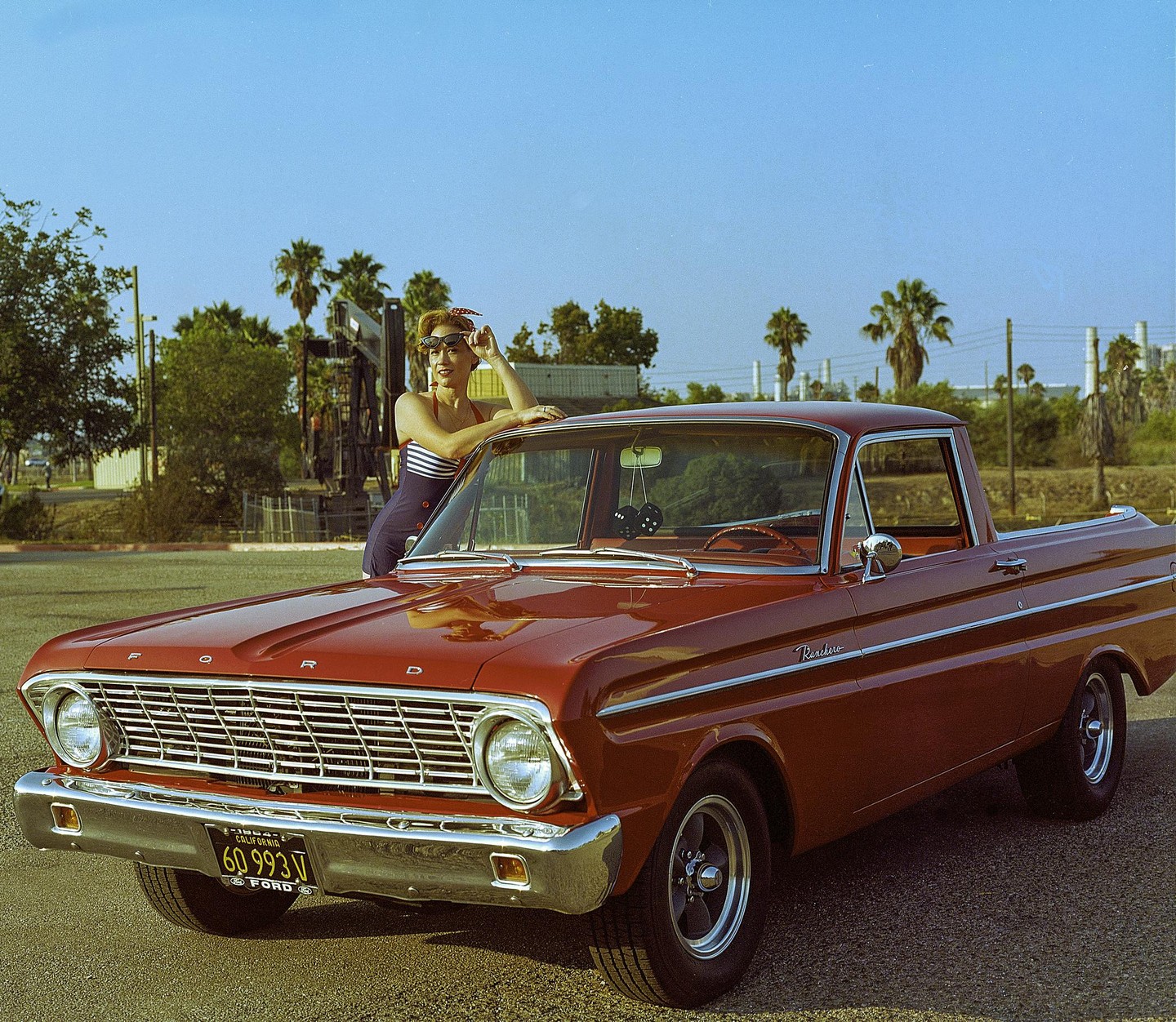 A woman in a striped swimsuit leans on a red vintage car with palm trees and industrial structures in the background.