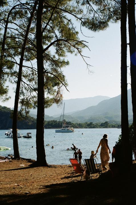 A woman and child walk towards the water on a sunny beach with boats and mountains in the background.