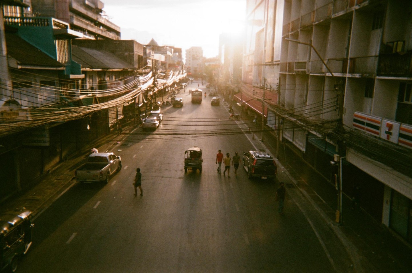 A street in Bangkok is filled with cars, tuk-tuks, and pedestrians under a bright sunset.