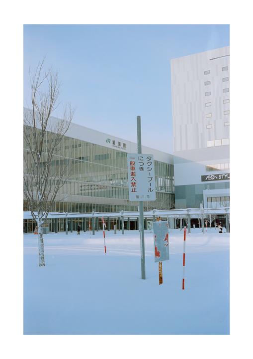 A snow-covered plaza with a bare tree and buildings in the background is seen under a clear sky.