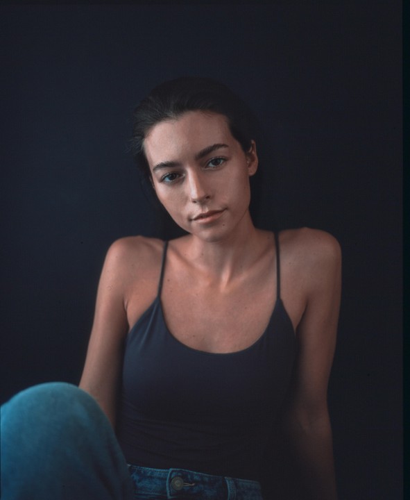 A woman in a dark tank top and jeans sits against a dark background.
