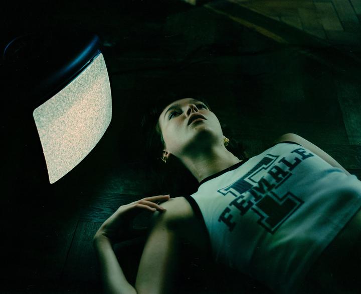 A woman lies on a dark floor, looking up at a television screen displaying static.