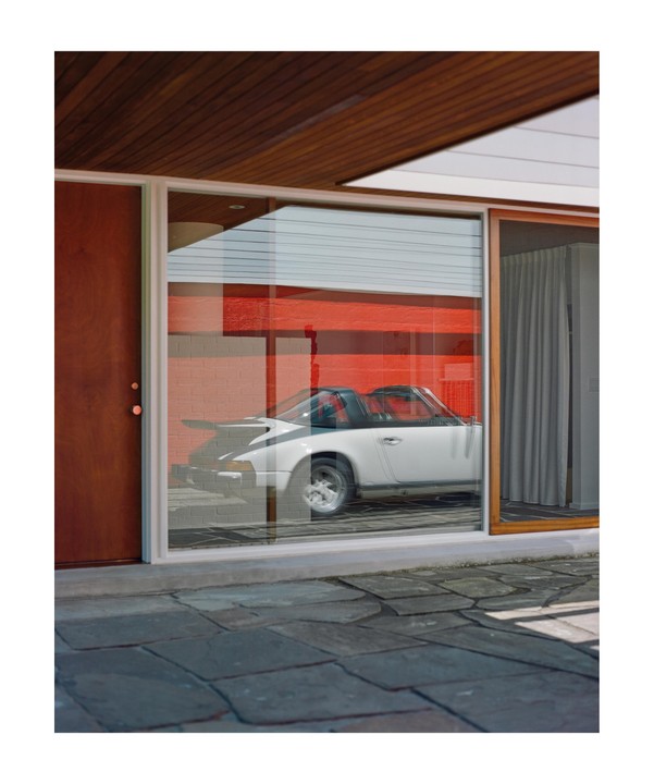 A white car is parked inside a building with a large window, reflecting a red wall.