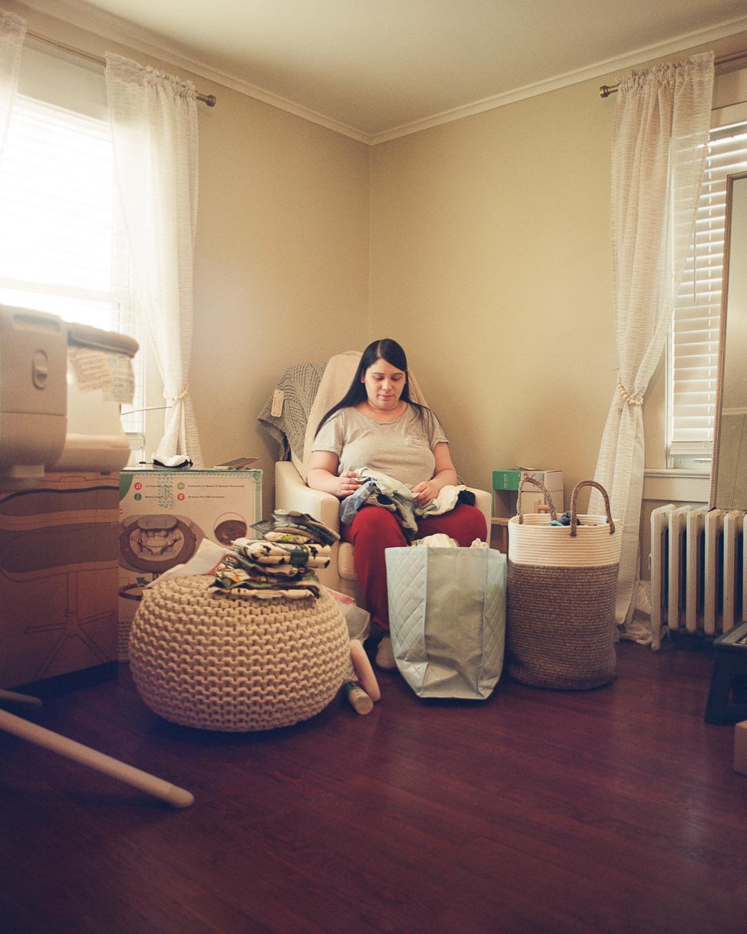 A woman sits in a chair folding clothes in a room with hardwood floors and two windows.