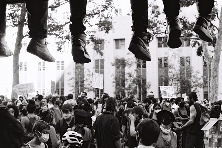 Several pairs of legs dangle above a crowd of people at a Black Lives Matter protest.