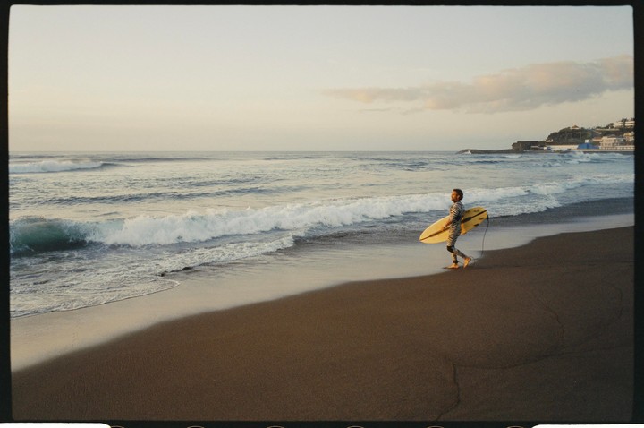 A man in a striped wetsuit walks barefoot on a dark sand beach carrying a surfboard as waves crash on the shore.