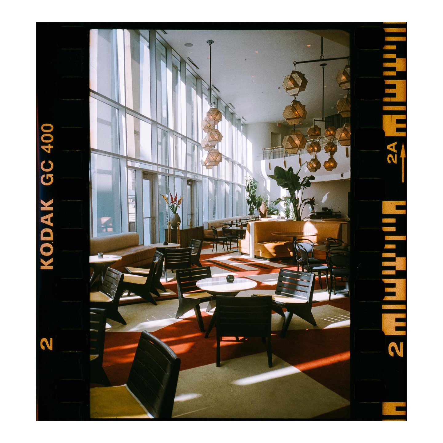Sunlight streams into a hotel lobby with tables, chairs, and hanging light fixtures.