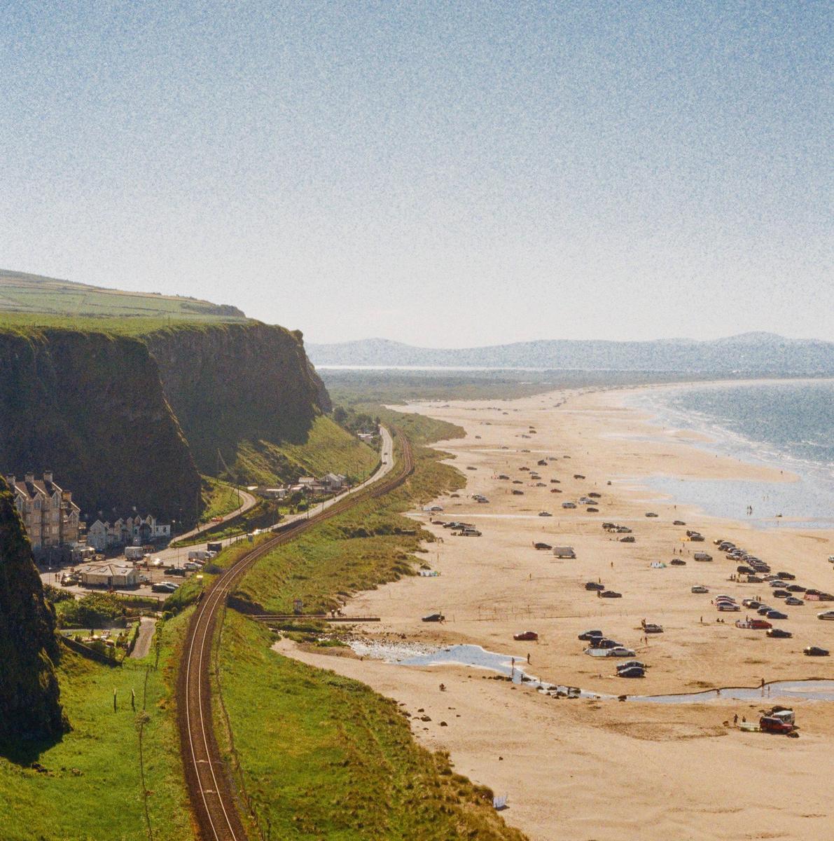 A long stretch of sandy beach with many cars parked on it is visible next to a railway line and a cliff.