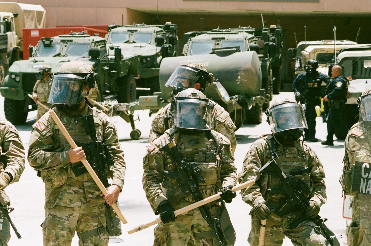 Several soldiers in camouflage and riot gear stand in front of military vehicles, some holding batons.