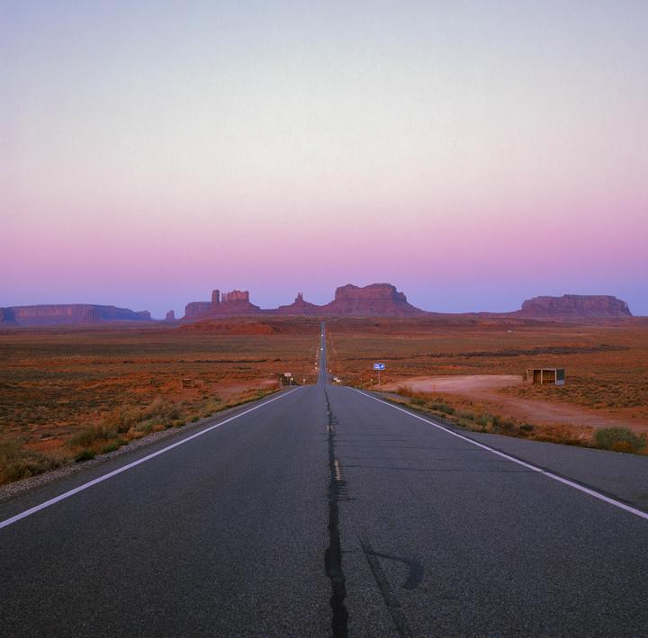A long, straight road leads towards distant mesas and buttes under a colorful sky at dusk.