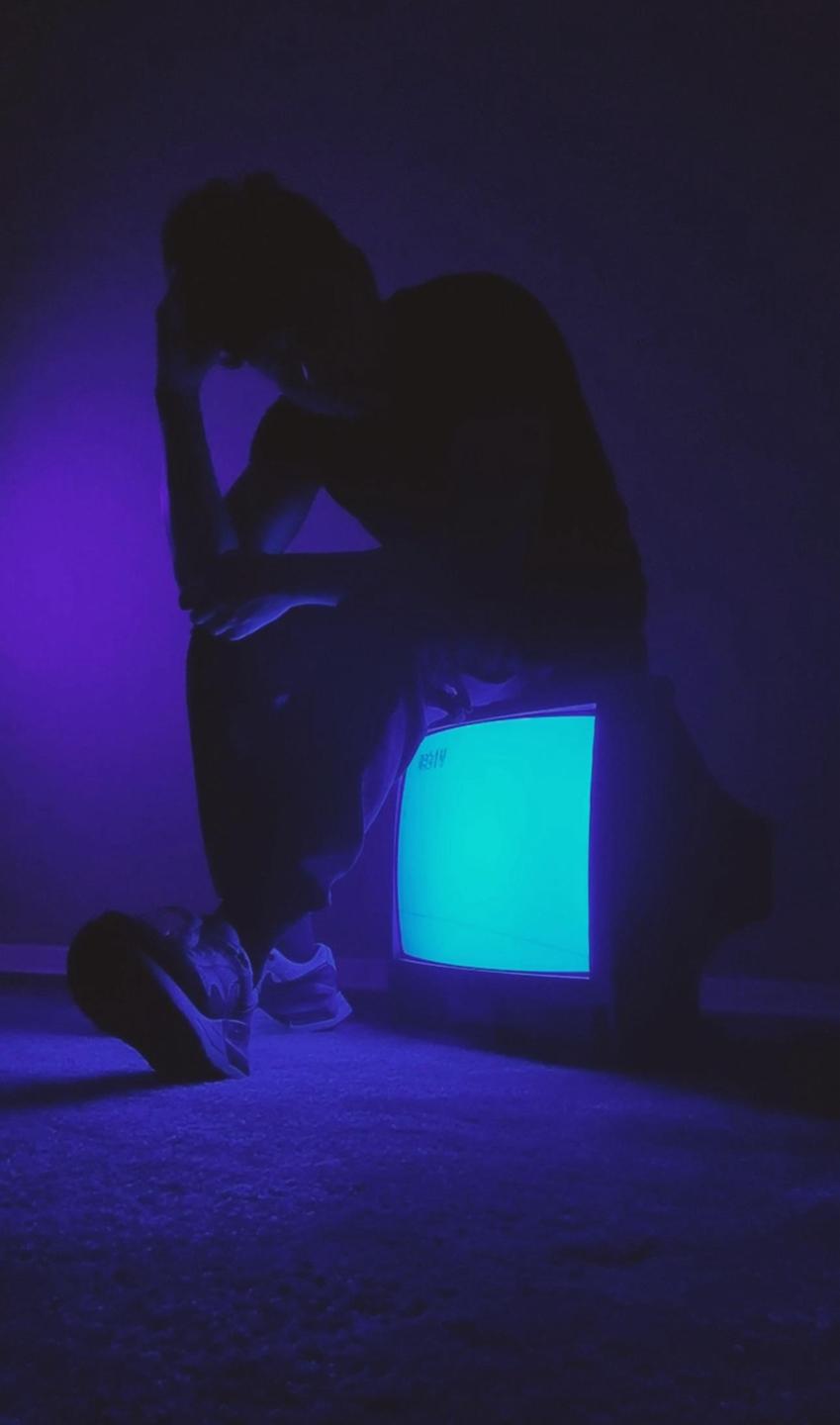 A man is silhouetted against a purple wall, illuminated by the glow of a television screen.